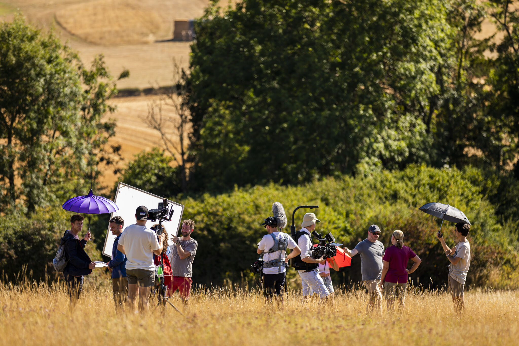 Film crew with camera and sound equipment preparing a scene in a sunny field with trees in the background.