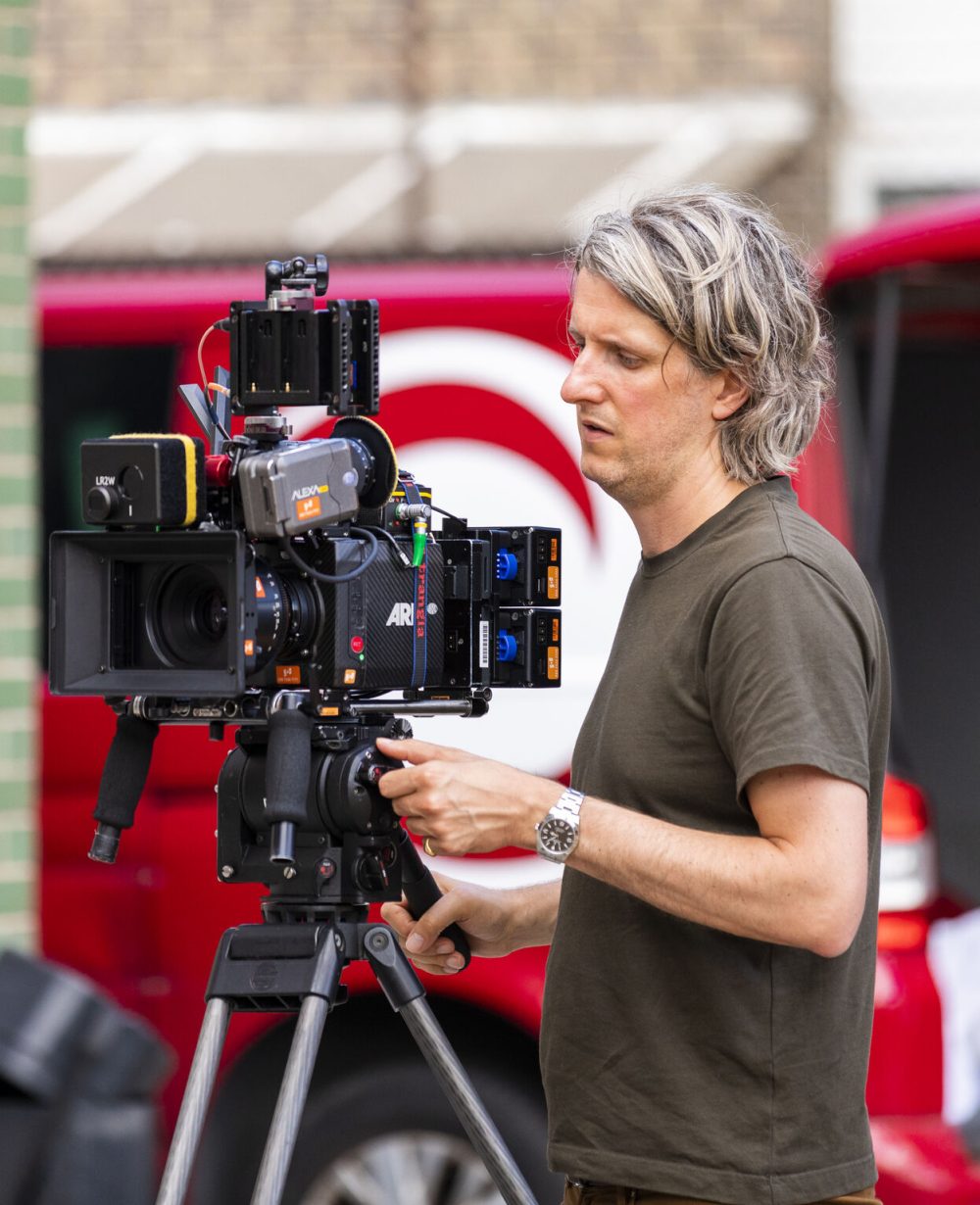 Focused man operating an ARRI camera on a tripod, with an urban backdrop and a red van, outdoors.
